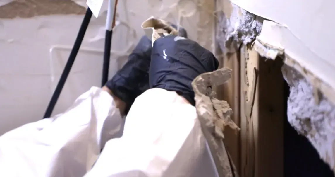 Technician in a protective suit removing moldy drywall and insulation inside a contained work area